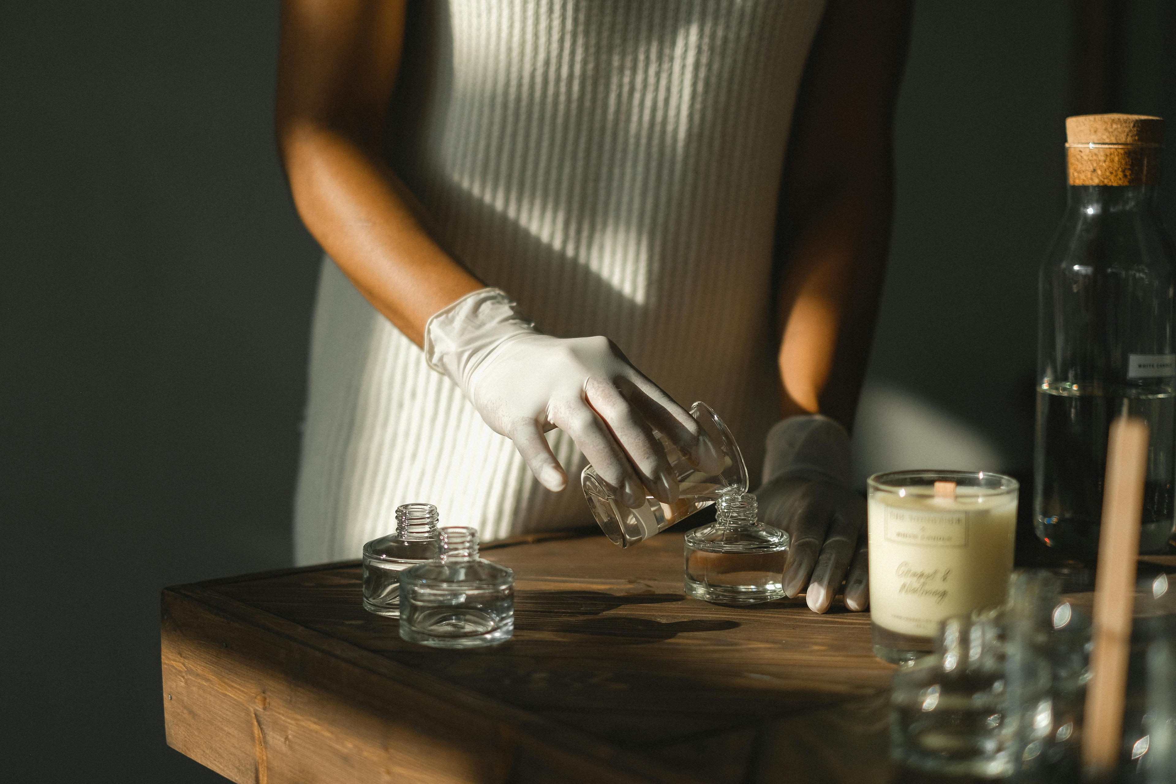 Person wearing gloves handling small glass containers on a wooden surface with bottles and a candle in the background.